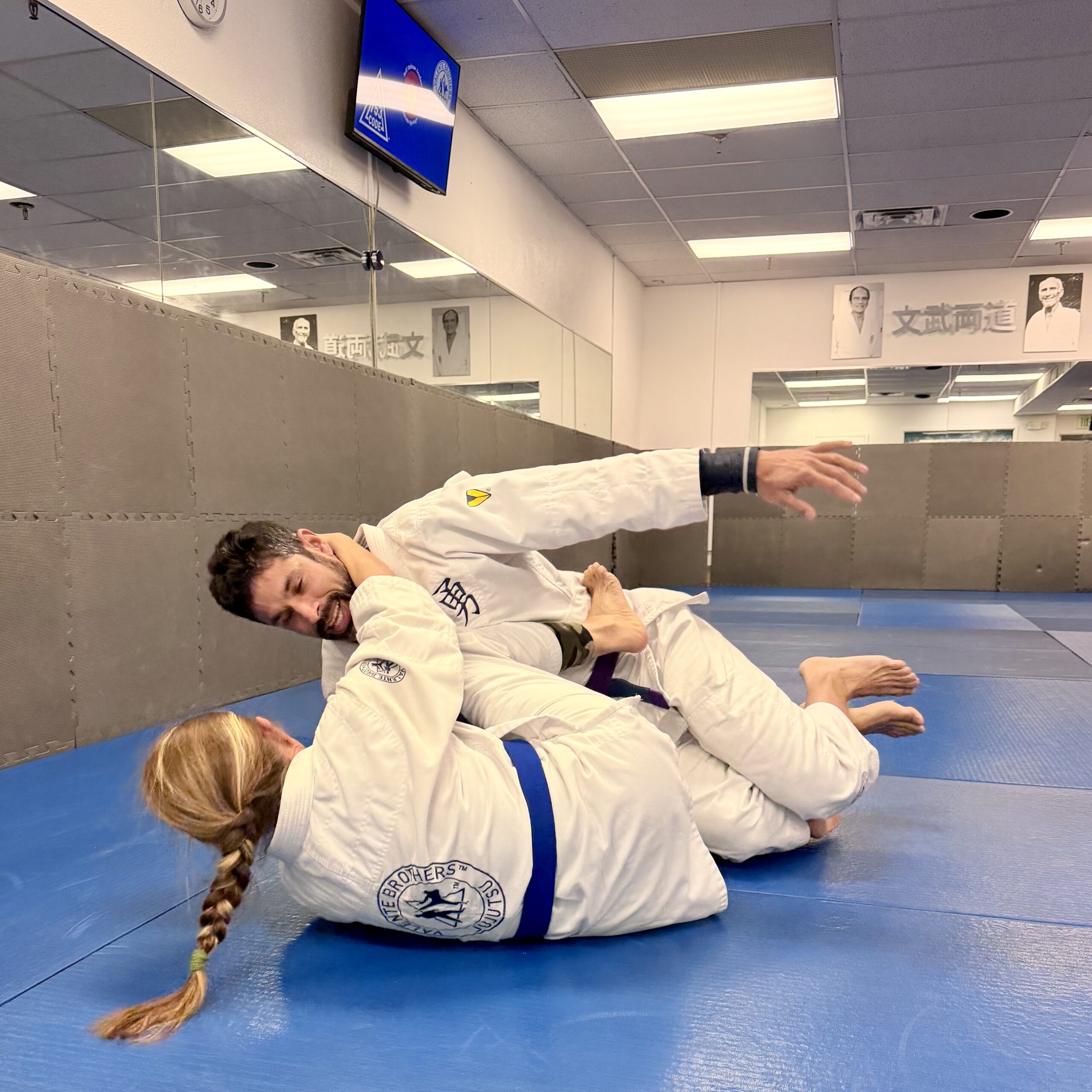 Two individuals in white martial arts kimonos practice Jiu-Jitsu on blue mats in a dojo. Mirrors and posters are visible in the background.
