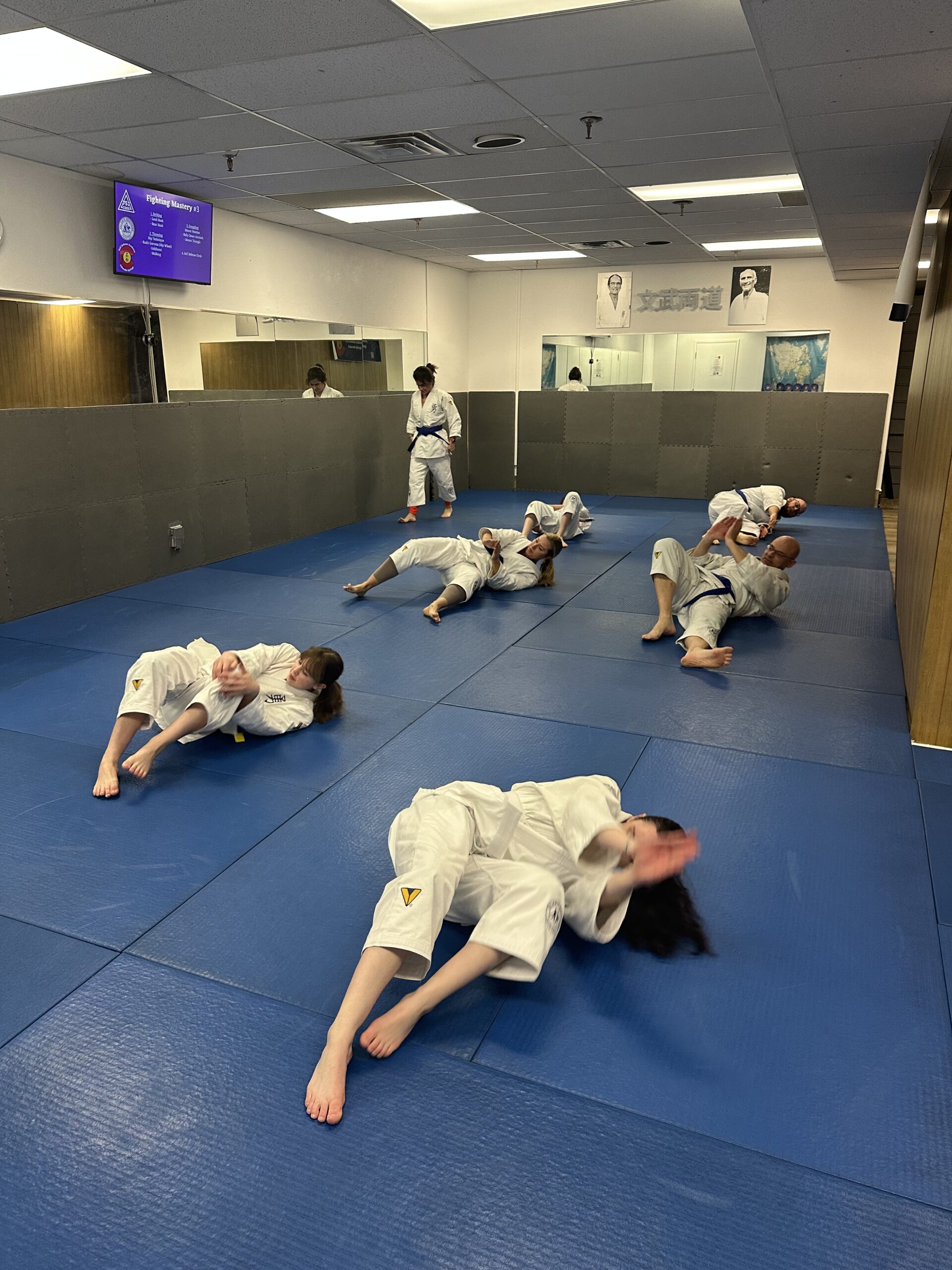 People practice martial arts rolls on blue mats in a dojo with a mirrored wall. A purple sign above reads "Fighting Mastery 1."