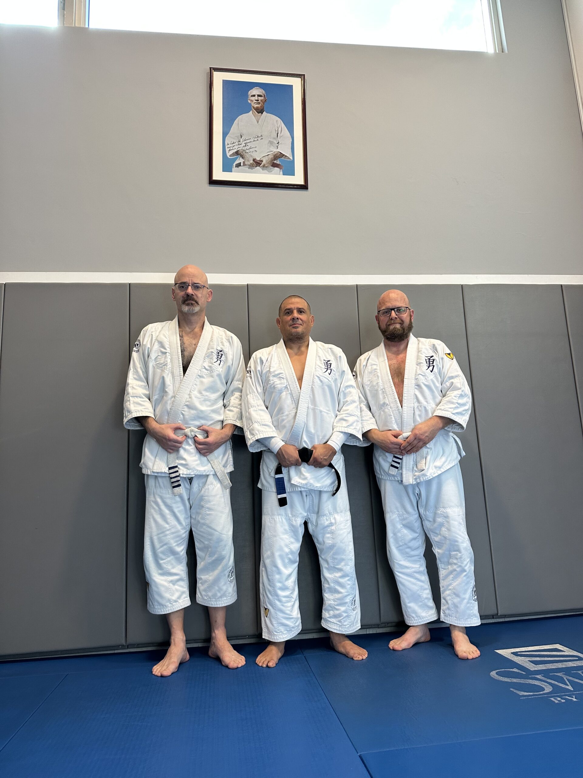 Martial arts class in session. Five people in white uniforms practice on blue mats. Mirrors reflect the scene. Posters on walls enhance focus.
