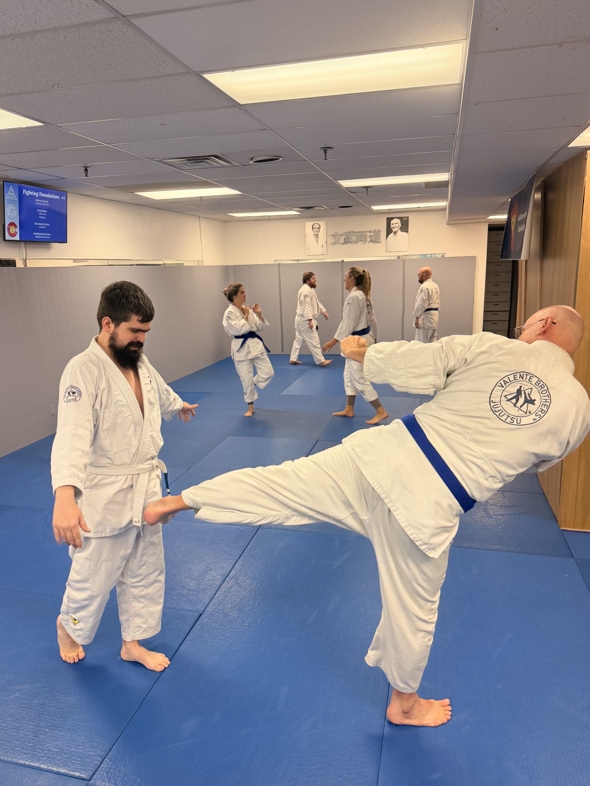 Adult jiu-jitsu class in white gis on blue mats; one man kicks another while others practice in a bright dojo with Valente Brothers sign.