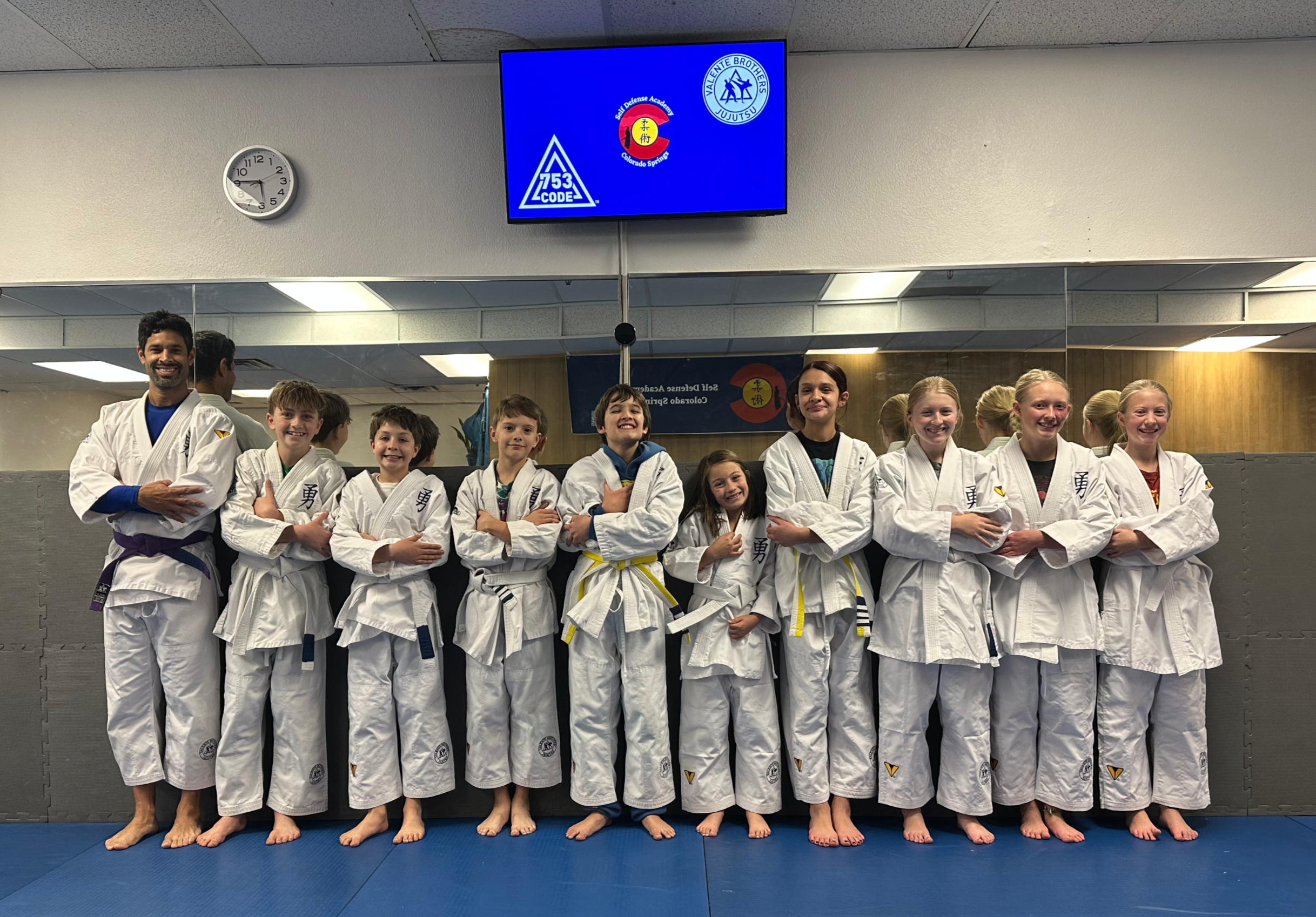 Junior jiu jitsu students in white gis posing with their instructor beneath the 753 Code and Valente Brothers logos at Self Defense Academy Colorado Springs